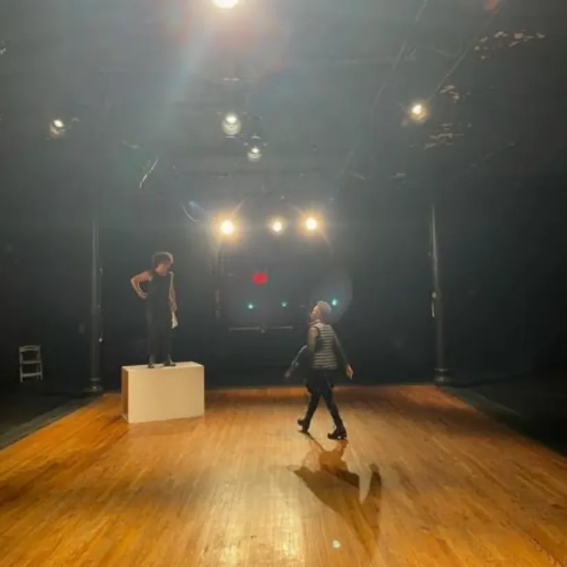 Production photo — an actor standing on a white wooden box under the lighting rig while another crosses the stage floor
