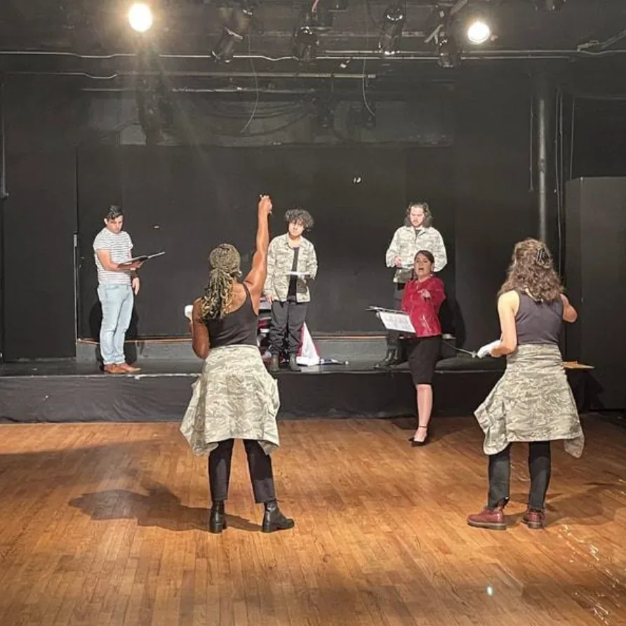 Production photo — six cast members holding scripts across the stage during the semi-staged reading