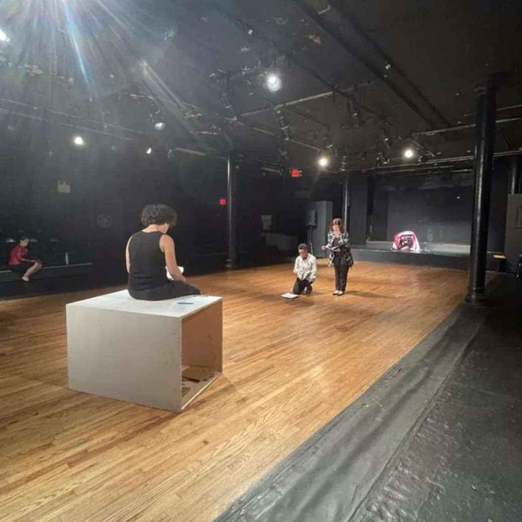 Production photo — cast members working on the wooden stage floor at The Flamboyán Theatre, one actor seated on a white wooden box and another kneeling over script pages